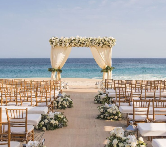 Beach wedding theme with a floral-draped arch facing the ocean, white and gold seating, and an elegant seaside ceremony setup.