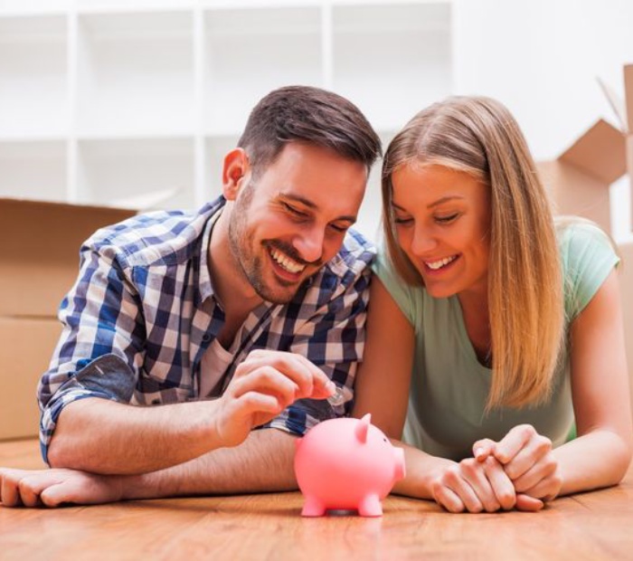 Couple smiling while placing a coin into a piggy bank at home, symbolizing saving money and building an emergency fund together.