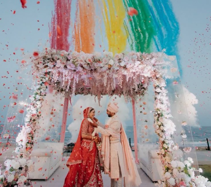 Destination wedding theme with a bride and groom under a floral mandap by the sea, colorful powder in the sky, and a scenic beach backdrop.