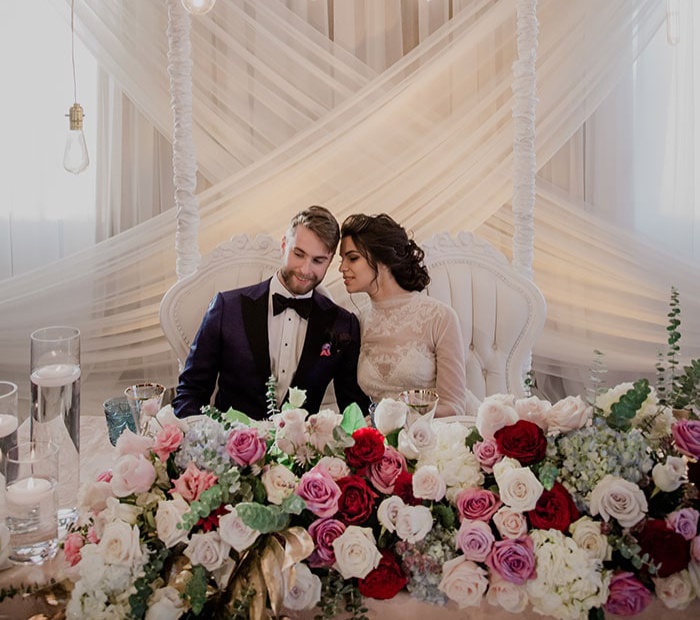 Modern wedding theme with an elegant bride and groom seated at a floral-decorated table, soft neutral drapes, and a stylish contemporary setup.