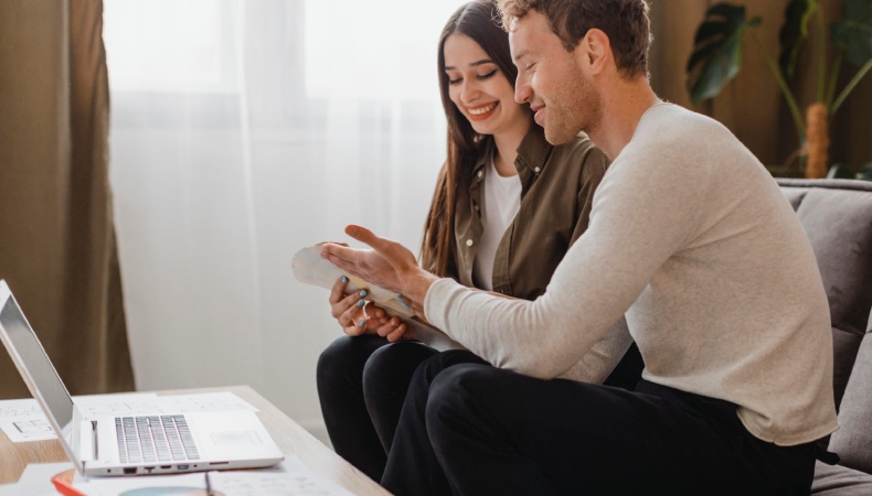 Newlywed couple sitting together at home reviewing financial documents and a laptop, smiling while planning their budget and future finances together.