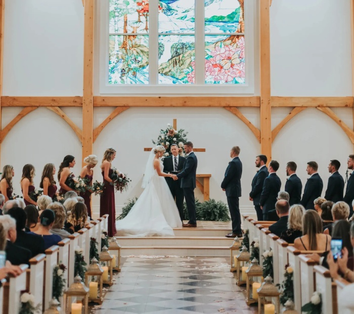 Traditional religious wedding ceremony inside a church with bride and groom at the altar, bridal party standing beside them, and guests seated along a decorated aisle.
