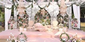 Elegant outdoor wedding venue with floral stage décor, crystal chandeliers, pastel seating, and a romantic garden wedding theme.