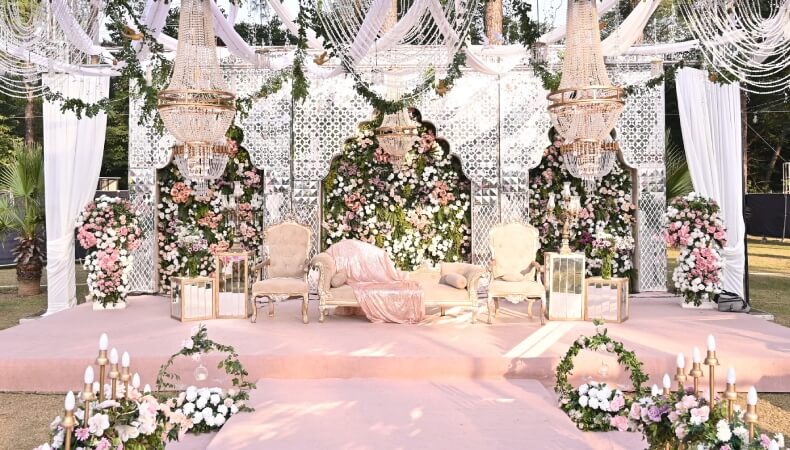 Elegant outdoor wedding venue with floral stage décor, crystal chandeliers, pastel seating, and a romantic garden wedding theme.