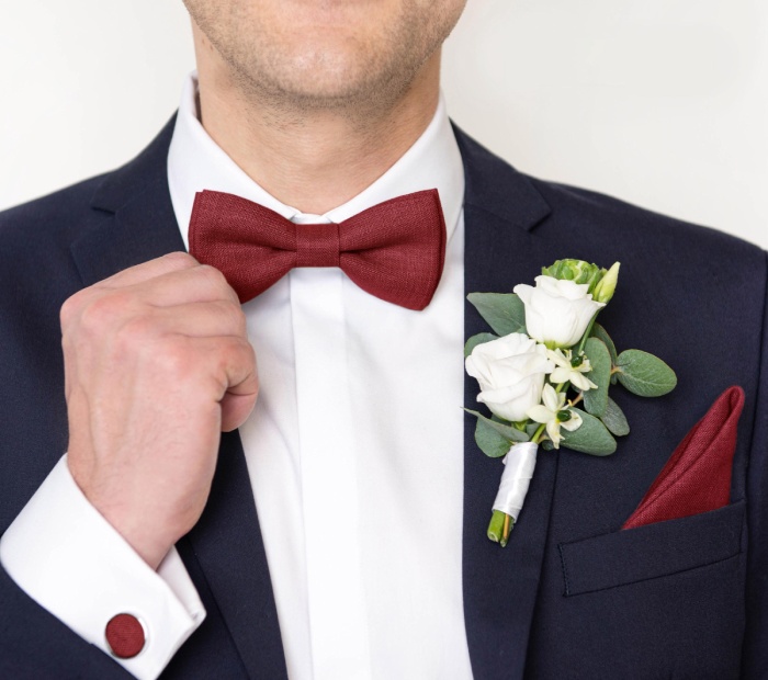 Close-up of a groom wearing a navy suit, white shirt, and burgundy bow tie with matching pocket square and cufflinks, holding the bow tie. A white floral boutonniere is pinned to the lapel.