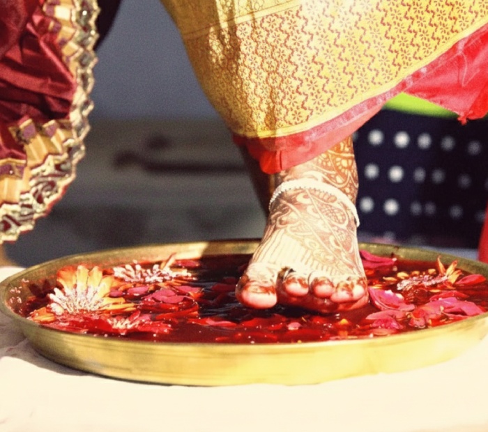A bride's foot, decorated with henna and a silver anklet, steps into a golden plate filled with red water and flower petals.
