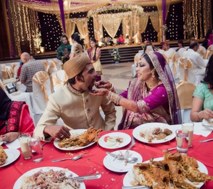 A bride in traditional attire playfully feeds her groom during a wedding feast at a large, decorated venue.