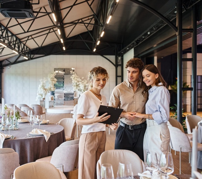 An event planner discusses a clipboard with a couple in a modern banquet hall, illustrating Capacity Planning – Number of Guests to Fit for the venue.