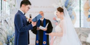 Groom reading wedding vows from a book while smiling bride listens during an indoor wedding ceremony with an officiant in the background.