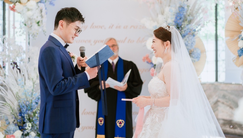 Groom reading wedding vows from a book while smiling bride listens during an indoor wedding ceremony with an officiant in the background.