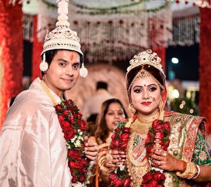 A Bengali bride in a red saree performing the Shubho Drishti ritual, holding betel leaves over her eyes with hands adorned in intricate henna and gold jewelry.
