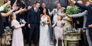 Bride and groom smiling as guests shower them with confetti outside a venue, with flower girls in pink dresses and family celebrating around them.