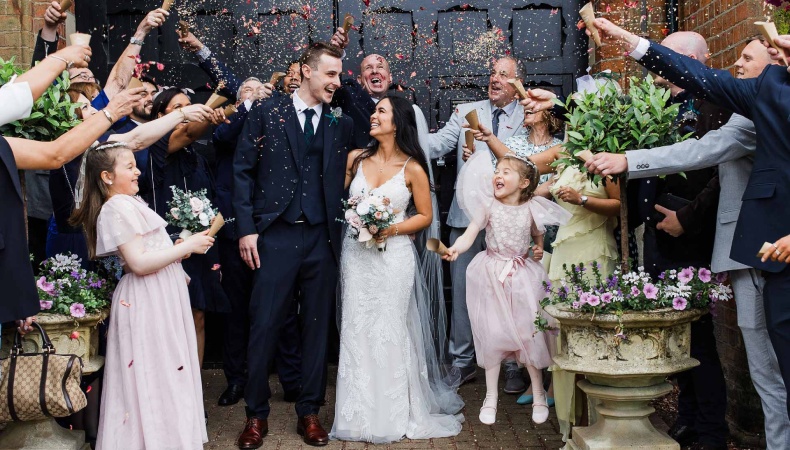 Bride and groom smiling as guests shower them with confetti outside a venue, with flower girls in pink dresses and family celebrating around them.