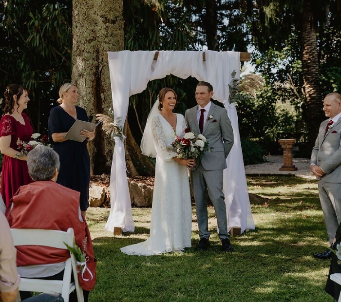 A bride and groom smiling under a white fabric wedding arch during an outdoor ceremony, with the officiant and bridal party nearby and guests seated on white chairs.