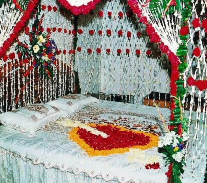 A bed decorated with white linens and red and yellow flower petals arranged in a heart shape, surrounded by elaborate floral garlands and curtains.