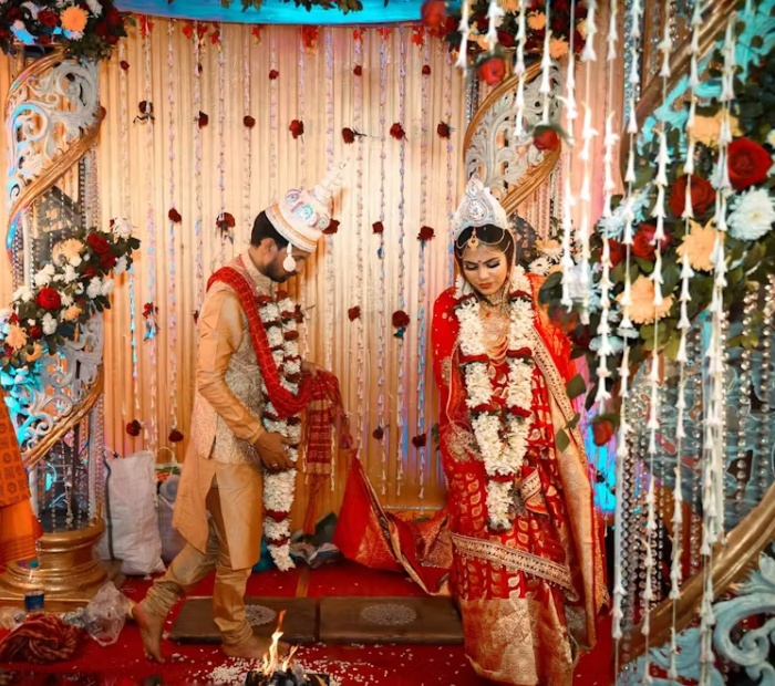 A smiling Bengali bride and groom in traditional wedding attire, wearing white Topor headpieces and red rose garlands, standing together during their ceremony.