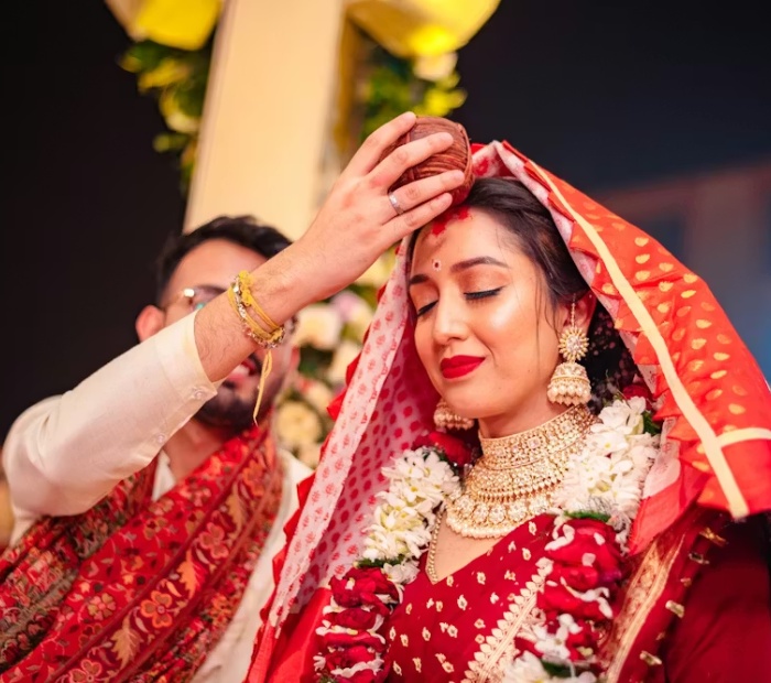 A smiling Bengali bride and groom in traditional wedding attire, wearing white Topor headpieces and red rose garlands, standing together during their ceremony.