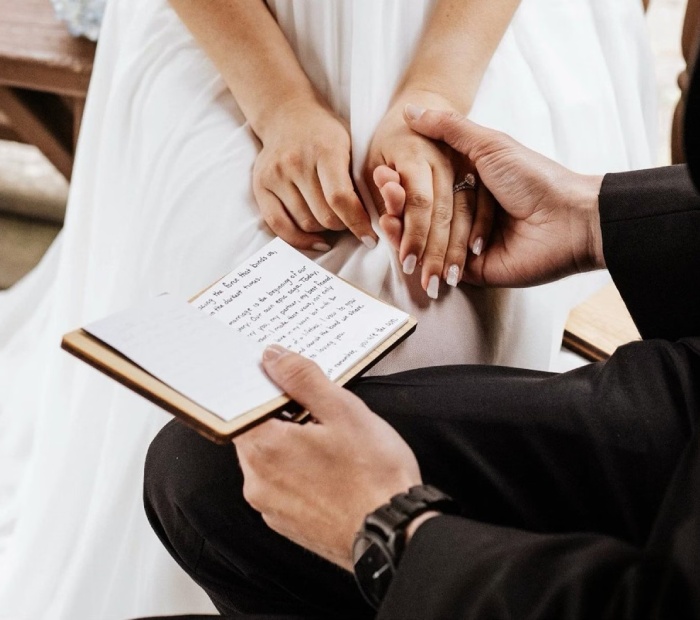 Close-up of couple holding hands as groom reads handwritten wedding vows from a notebook during an intimate ceremony.