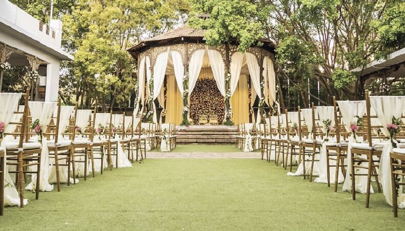 An outdoor wedding venue with a decorated wooden gazebo as a central altar, flanked by rows of gold Tiffany chairs on a green lawn.