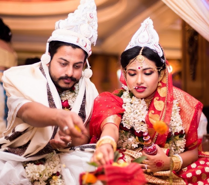 A smiling Bengali bride and groom in traditional wedding attire, wearing white Topor headpieces and red rose garlands, standing together during their ceremony.
