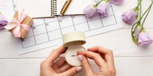 Hands holding an open ring box with an engagement ring on a white desk, with a calendar page, notebook, pen, gift box, and purple flowers in the background.