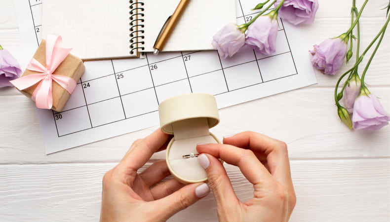 Hands holding an open ring box with an engagement ring on a white desk, with a calendar page, notebook, pen, gift box, and purple flowers in the background.