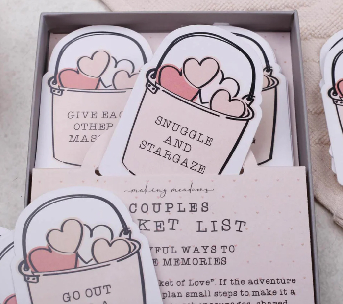 A box of "Couples Bucket List" cards featuring bucket-shaped illustrations filled with hearts, with activity prompts like "Snuggle and Stargaze" and "Go Out For A Karaoke."