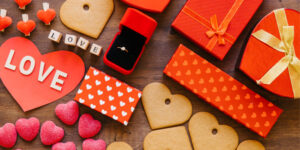An overhead shot of a wooden surface covered in Valentine's Day items, including heart-shaped cookies and candies, red gift boxes, a ring in a velvet box, and a paper heart with the word "LOVE."