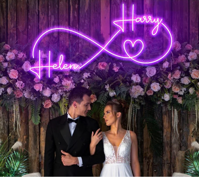 “Bride and groom posing beneath a purple neon infinity sign with floral wedding backdrop.”