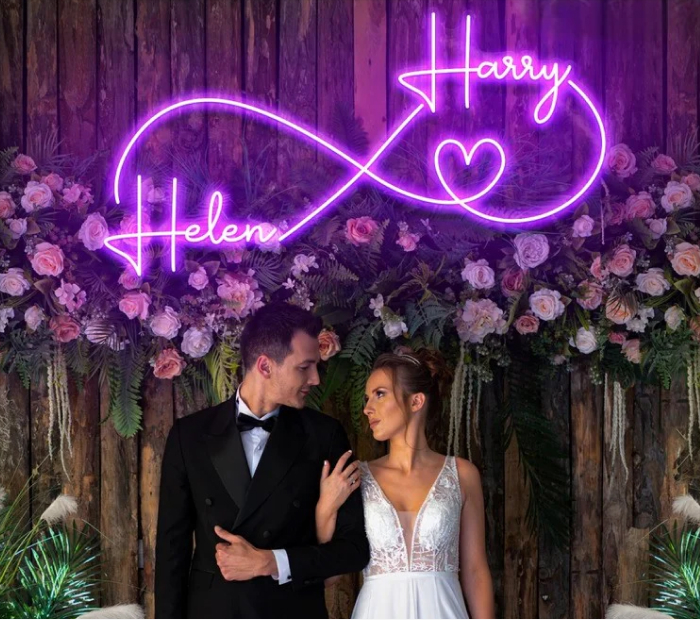 “Bride and groom posing beneath a purple neon infinity sign with floral wedding backdrop.”