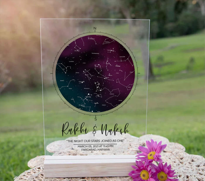 Personalized star map acrylic plaque with constellations and a couple’s names, placed on a table outdoors with pink flowers beside it.