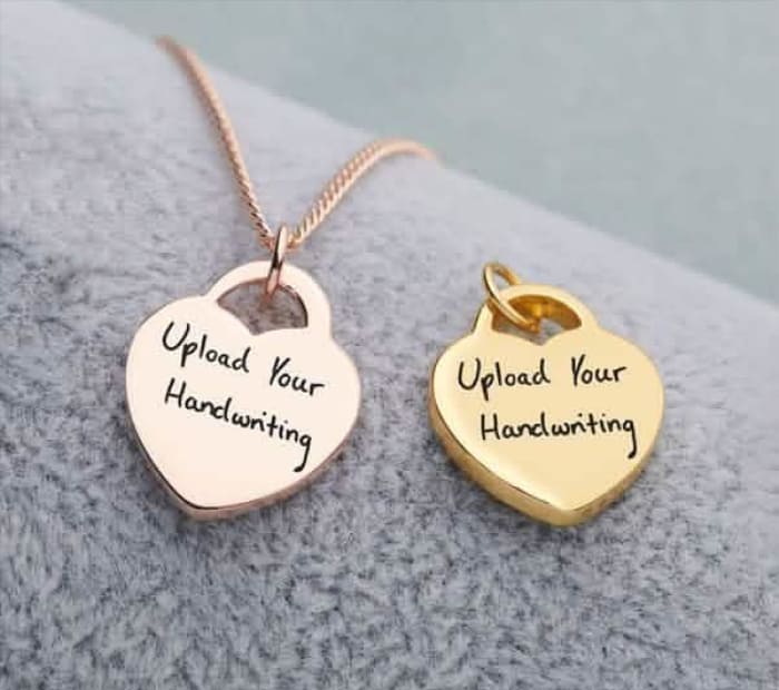 Two heart-shaped pendant charms in rose gold and gold engraved with “Upload Your Handwriting,” displayed on a soft fabric background.