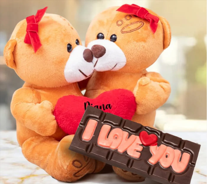 Two teddy bears with red bows sitting together and holding a plush red heart, positioned behind a chocolate bar that says "I Love You" in pink lettering.