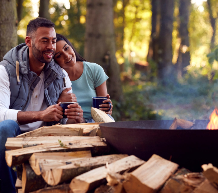 Couple relaxing by a campfire in the woods, holding warm drinks and enjoying a cozy outdoor moment, ideal for a romantic Valentine’s Day experience gift.