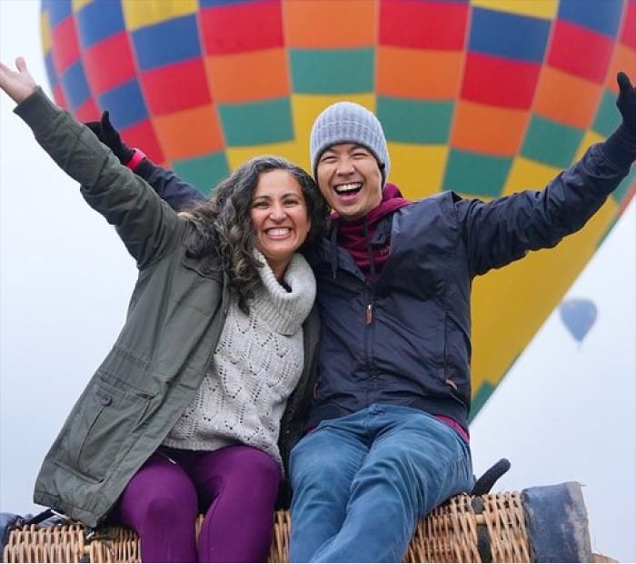 Joyful couple sitting in a hot air balloon basket with arms raised, smiling together during a romantic adventure experience.
