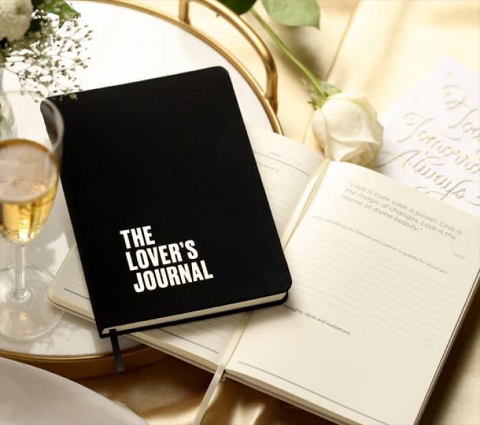 Romantic “The Lover’s Journal” notebook placed beside an open journal, white rose, and champagne glass on a tray.