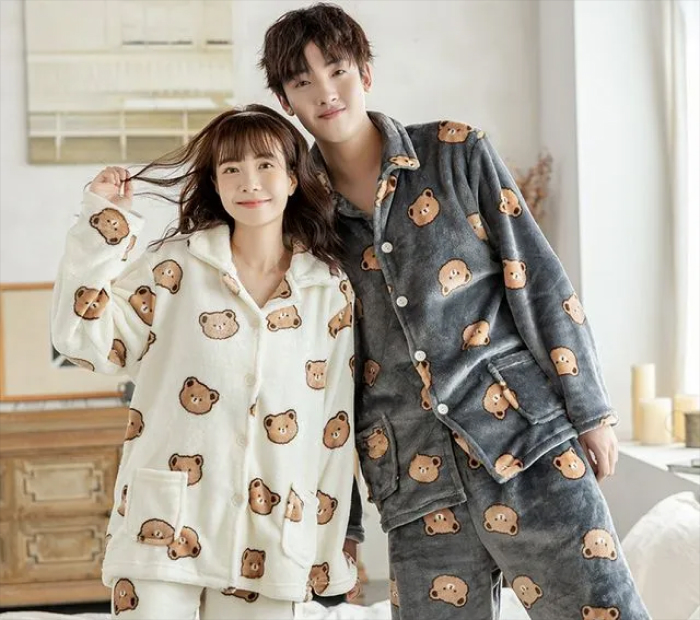 Smiling couple wearing matching teddy bear print pajamas, standing together in a cozy bedroom setting.