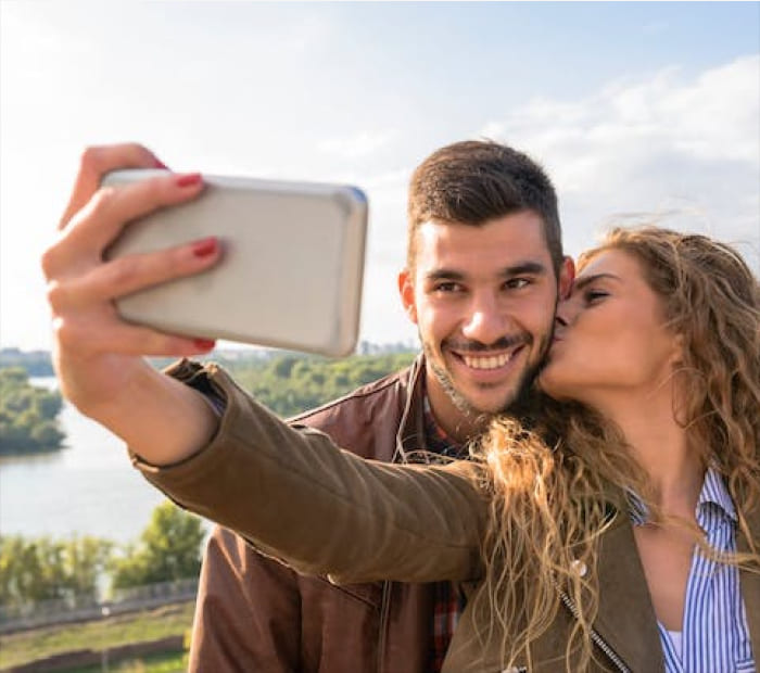 Happy couple taking a selfie outdoors by the river, with a woman kissing her partner, capturing a romantic Valentine’s Day moment.