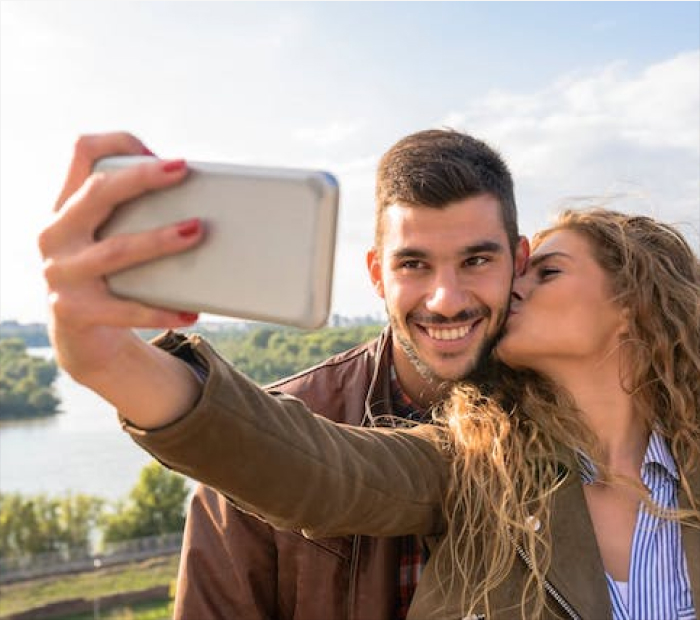Happy couple taking a selfie outdoors by the river, with a woman kissing her partner, capturing a romantic Valentine’s Day moment.