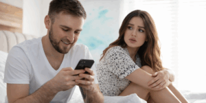 Man sitting on a bed smiling at his phone while a woman beside him looks at him with a worried and suspicious expression in a bright bedroom setting.