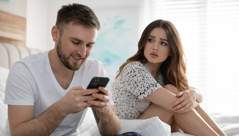 Man sitting on a bed smiling at his phone while a woman beside him looks at him with a worried and suspicious expression in a bright bedroom setting.