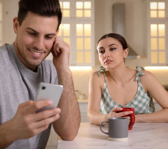 A man smiles while looking at his phone in a bright kitchen as a woman holding a red mug watches him with a concerned expression.