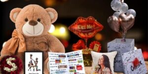 A Valentine’s Day gift setup with a teddy bear, heart-shaped balloons, a “Kiss Me” sign, roses, chocolates, a personalized photo pillow, and gift boxes on a wooden table with warm bokeh lights in the background.