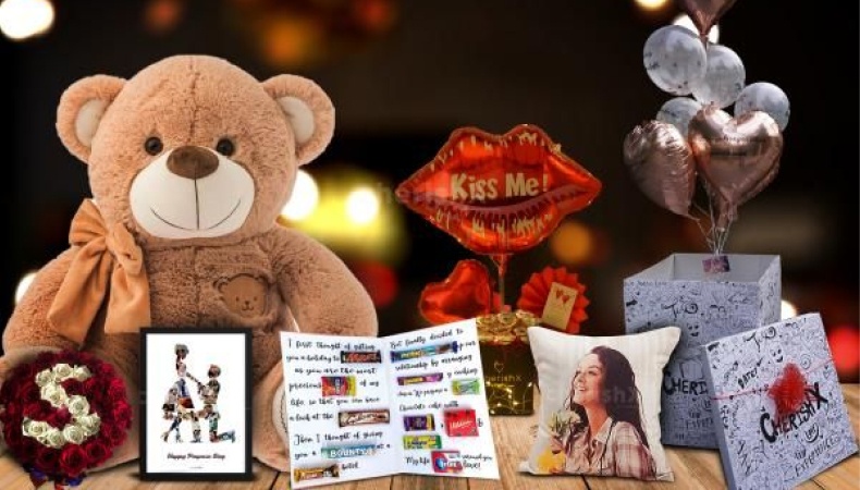 A Valentine’s Day gift setup with a teddy bear, heart-shaped balloons, a “Kiss Me” sign, roses, chocolates, a personalized photo pillow, and gift boxes on a wooden table with warm bokeh lights in the background.