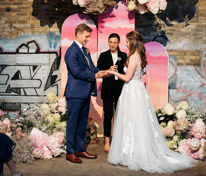 Bride and groom exchange rings beneath suspended floral arrangements during a modern indoor wedding ceremony.