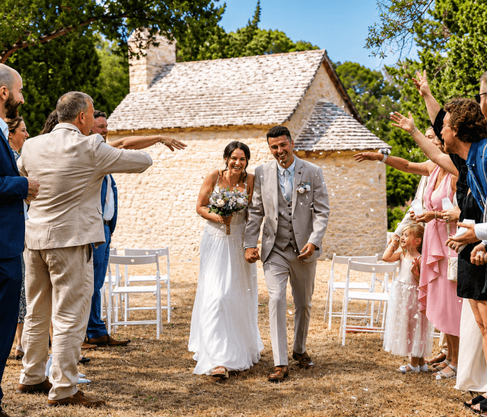 Happy newlywed couple walking through cheering wedding guests outside a rustic stone chapel during an outdoor ceremony.