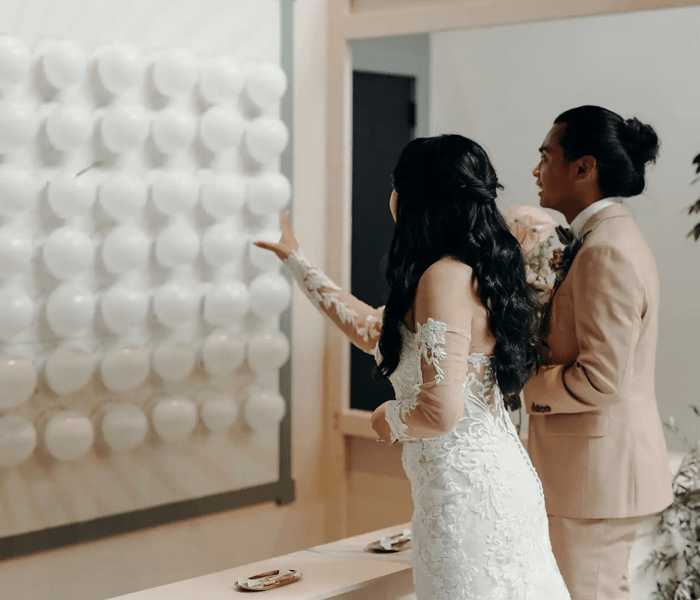 Balloon Dart Game - Happy Wedding App A bride in a lace wedding dress aiming a dart at a wall covered in white balloons.