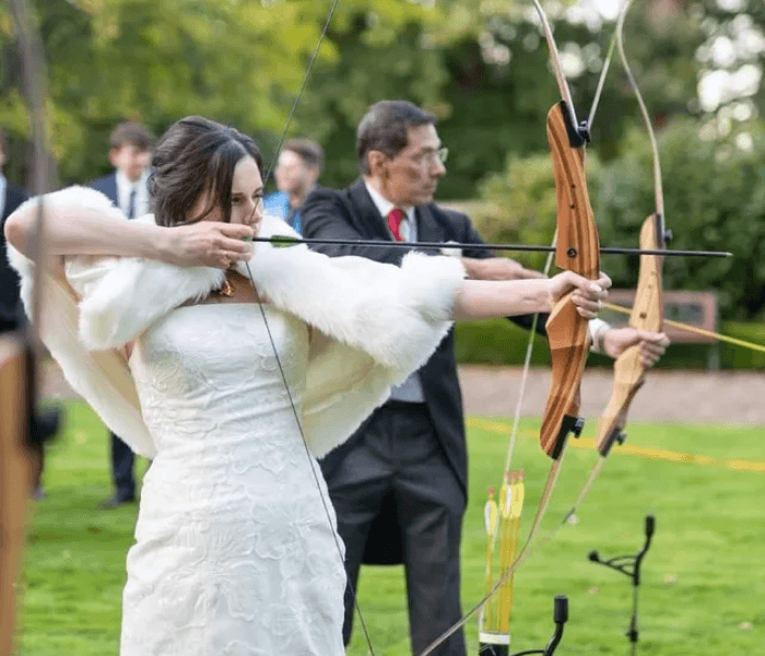 Foam-Tipped Archery Range - Happy Wedding App A bride in a white gown aims a wooden bow and arrow outdoors alongside a man.