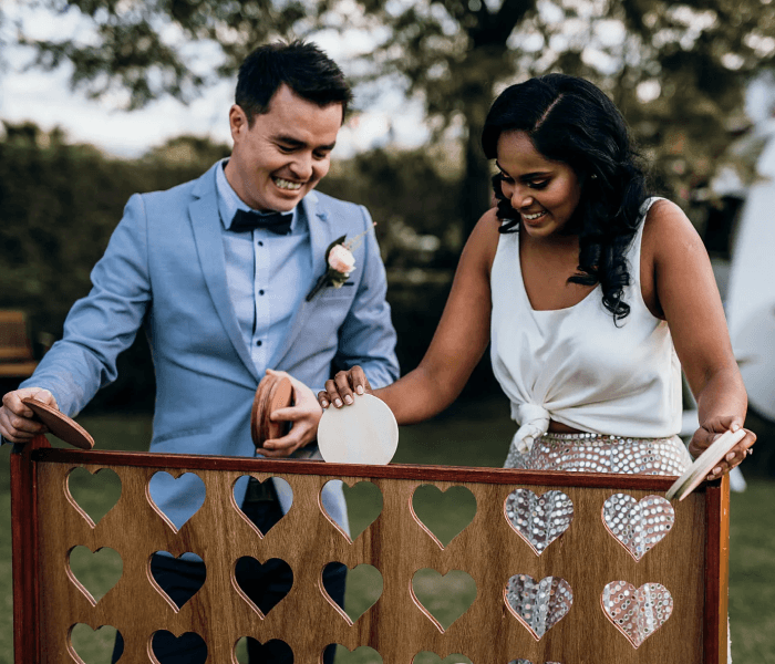 Giant Connect Four - Happy Wedding App A man and woman dropping wooden discs into a large wooden heart-cutout Connect Four game outdoors.