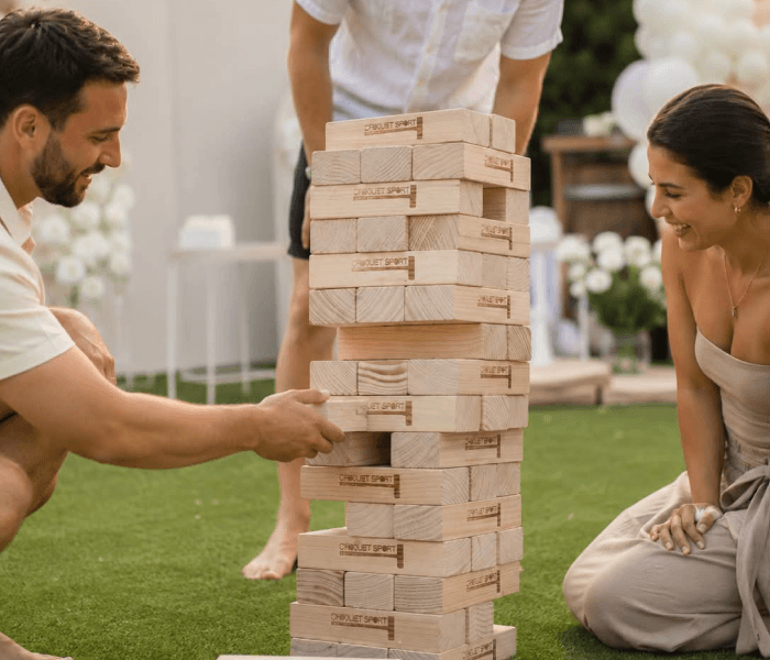 Giant Jenga - Happy Wedding App A man and woman dropping wooden discs into a large wooden heart-cutout Connect Four game outdoors.